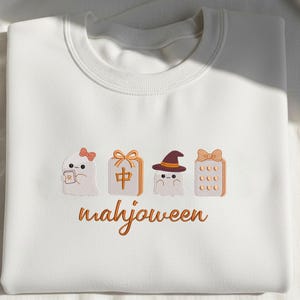May include: A white crewneck sweatshirt featuring embroidered designs of two ghosts, a mahjong tile, and a mahjong rack, all accented with orange bows and a witch hat. The text "mahjoween" is embroidered in orange cursive script below the icons.