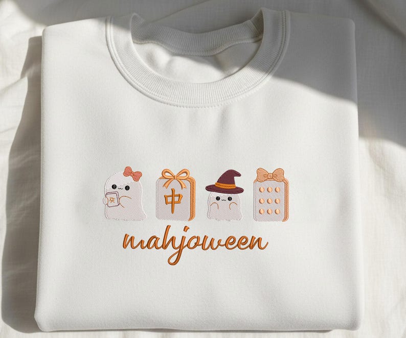 May include: A white crewneck sweatshirt featuring embroidered designs of two ghosts, a mahjong tile, and a mahjong rack, all accented with orange bows and a witch hat. The text "mahjoween" is embroidered in orange cursive script below the icons.