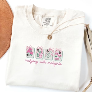May include: Cream-colored t-shirt with embroidered mahjong tiles in pink, green, and white. The text "mahjong with mahgirls" is embroidered below the tiles. A gold necklace and a brown hat are visible.