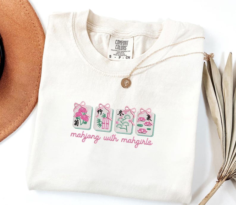 May include: Cream-colored t-shirt with embroidered mahjong tiles in pink, green, and white. The text "mahjong with mahgirls" is embroidered below the tiles. A gold necklace and a brown hat are visible.