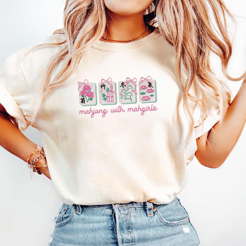 May include: Cream-colored t-shirt featuring a graphic of four embroidered Mahjong tiles with pink bows and floral designs. The text "mahjong with mahgirls" is written in pink script below the tiles. The shirt is paired with blue jeans.