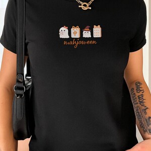 May include: A black short-sleeved t-shirt featuring a graphic print of two ghosts and two mahjong tiles. One ghost wears a pink bow, the other a witch hat. The tiles include a red character and a dotted pattern. The text "mahjoween" is printed below.