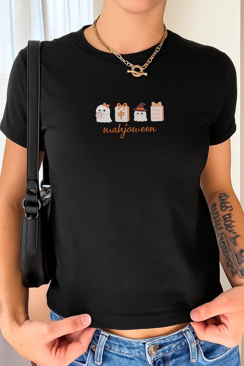 May include: A black short-sleeved t-shirt featuring a graphic print of two ghosts and two mahjong tiles. One ghost wears a pink bow, the other a witch hat. The tiles include a red character and a dotted pattern. The text "mahjoween" is printed below.