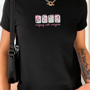 May include: Black t-shirt with a graphic featuring four Mahjong tile designs in pink, green, and white, with the text "mahjong with mahgirls" in a cursive font. The shirt is a fitted style.
