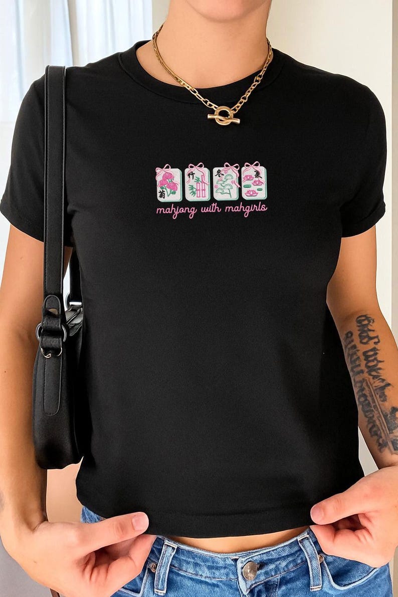 May include: Black t-shirt with a graphic featuring four Mahjong tile designs in pink, green, and white, with the text "mahjong with mahgirls" in a cursive font. The shirt is a fitted style.