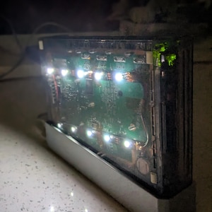 May include: A rectangular electronic device with a clear plastic cover, revealing a green circuit board and bright white LED lights. The device sits in a silver metal base, with a dark background.