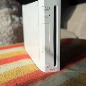 May include: A white Wii games console with power and reset buttons. The console has a slot for game discs and the word "Wii" printed on the front. The console is on a striped blanket.