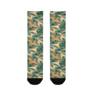 Puede incluir: Un par de calcetines con un patrón repetido de pterodáctilos naranjas y crema, árboles verdes y montañas sobre un fondo verde azulado. Los calcetines tienen punteras negras y están diseñados para la comodidad y el estilo.