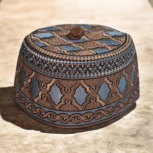 May include: A blue and brown embroidered skullcap with a geometric pattern. The cap features a brown top button and intricate stitching details. The design includes diamond and floral shapes.