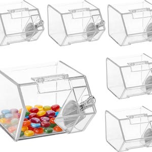 Puede incluir: Un conjunto de seis recipientes transparentes y angulados para caramelos. Un recipiente está lleno de caramelos de colores. Cada recipiente tiene una tapa con bisagras y un mecanismo dispensador. Los recipientes son de plástico transparente.