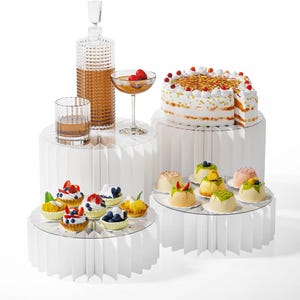 Op de afbeelding: Een display van desserts en drankjes op witte sokkels. Inclusief een taart, mini-taartjes met bessen en individuele puddings. Een karaf, een glas whisky en een cocktailglas zijn ook aanwezig. De sokkels hebben een geplooid ontwerp.