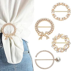 Puede incluir: Anillos y cierres de bufanda dorados con varios diseños. Uno está adornado con un lazo, mientras que otros presentan detalles de perlas y cristales. Los cierres se muestran en una parte superior blanca y jeans, mostrando su uso como accesorios de moda.