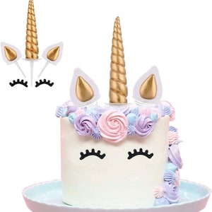 Puede incluir: Un pastel blanco decorado como un unicornio, con cuerno dorado, orejas y pestañas negras. El pastel está adornado con flores de glaseado de colores pastel y se encuentra sobre un plato azul claro. Se incluyen decoraciones adicionales de cuerno y oreja de unicornio.