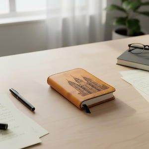 May include: A tan leather-bound notebook featuring a detailed dark brown laser-engraved illustration of a cathedral on the front cover, accompanied by a black ribbon bookmark and white pages, positioned alongside a black pen and a closed book.