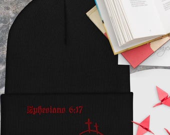 Puede incluir: Gorro negro con texto bordado en rojo "Efesios 6:17" y un diseño de cruz. El gorro está hecho de un material de punto suave. El borde está doblado. Un libro y origami en el fondo.