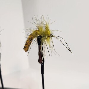 May include: Close-up of three artificial fishing flies. The flies are mounted on black stands against a white background. The flies are various colors, including olive green, tan, and gray, with feathery details and small hooks.