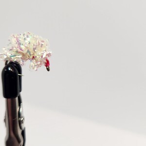 May include: A close-up of a small, decorative fly fishing lure. The lure features a cluster of iridescent, white, and pink materials, with a small red accent. It is attached to a black hook and held by a silver clamp.