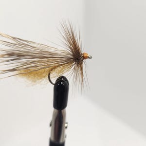 May include: A handmade fishing fly, close up. The fly has a hook with a brown and tan body, and brown and tan feather detailing. It is held in a black clamp against a white backdrop.