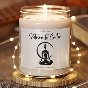 May include: A clear glass jar candle with a lit wick and light beige wax. The white label features the text Zen Flow Designs, Return to Center, and Eco-Friendly 9 ounce Soy Candle, along with a black graphic of a person in a seated yoga pose.