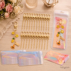 May include: A set of gold-toned metal lobster clasp keychains with attached chains, featuring charms shaped like a pineapple, a candy, and a hat, along with round letter charms K and D. Includes iridescent plastic pouches and printed thank you cards.