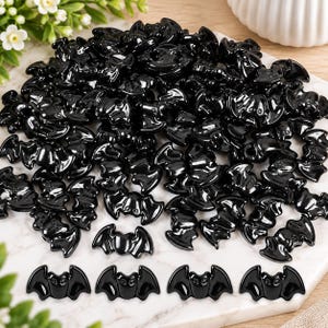 May include: A large collection of small, glossy black bat-shaped beads. Each bead features a stylized bat silhouette with defined wings and a central body. The beads are scattered in a pile, with four individual bat beads arranged in a row at the bottom.