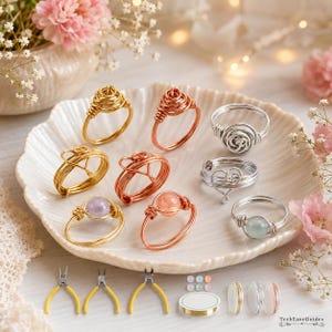 May include: A collection of wire-wrapped rings in gold, copper, and silver tones, some featuring round pastel beads and heart-shaped wire designs. The set includes three small pliers, a spool of wire, and loose beads arranged on a white shell dish.