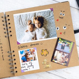 May include: Open scrapbook with photos of children and family events. The scrapbook has a spiral binding and a brown cover. The photos are secured with purple photo corners. Text includes "Sisters forever" and "Let's try this yummy cake!"