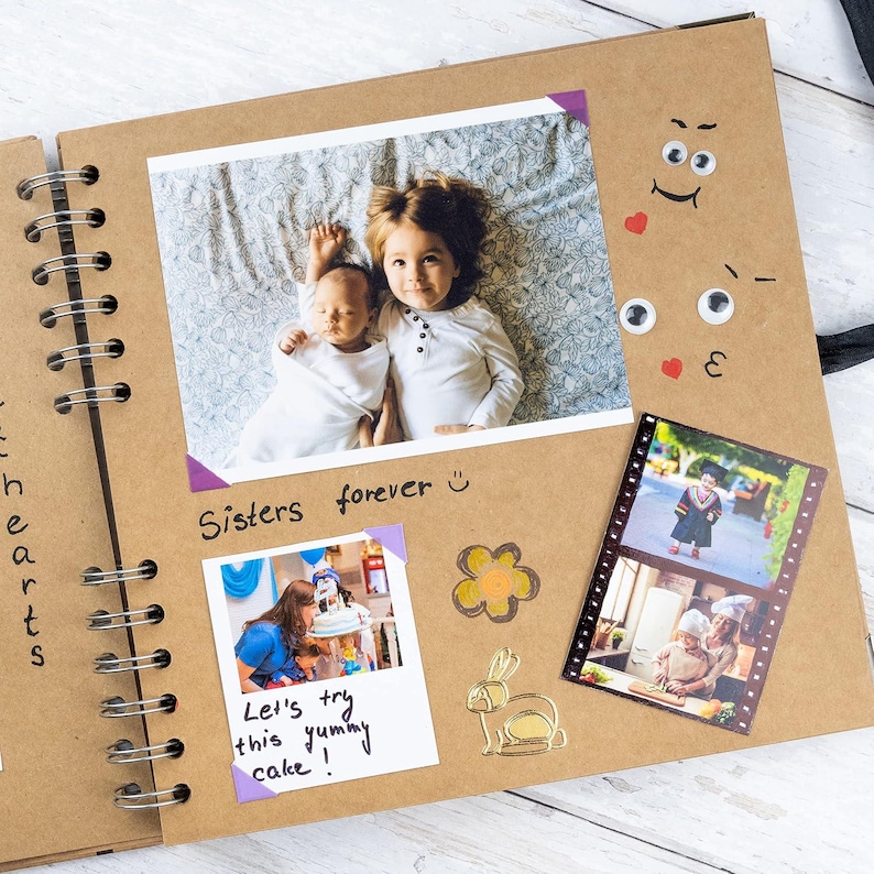 May include: Open scrapbook with photos of children and family events. The scrapbook has a spiral binding and a brown cover. The photos are secured with purple photo corners. Text includes "Sisters forever" and "Let's try this yummy cake!"