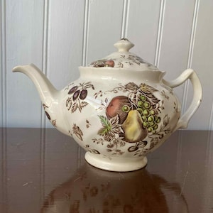 Peut inclure: Théière en céramique crème avec couvercle, ornée d'un motif de fruits dans les tons marron, vert et violet. La théière a un bec et une anse courbés. Le motif comprend des raisins, des poires et d'autres fruits.