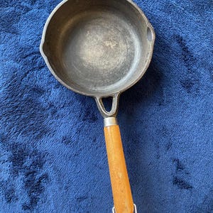 May include: A small cast iron frying pan with a wooden handle. The pan is dark grey with a round cooking surface and a single handle. The handle is light brown and attached with a metal ring. The skillet is designed for cooking.
