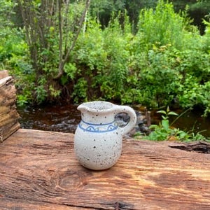 May include: A small ceramic pitcher with a speckled off-white glaze and a single handle. The pitcher features a hand-painted blue line pattern with small semi-circular arches encircling the upper neck area, providing a simple decorative detail.