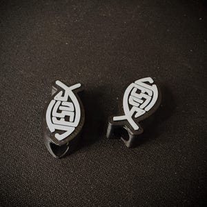 May include: Two black and white Christian fish symbols, each with the word "JESUS" inside. The fish symbols are on a dark surface. The symbols are likely pins or small decorative items.