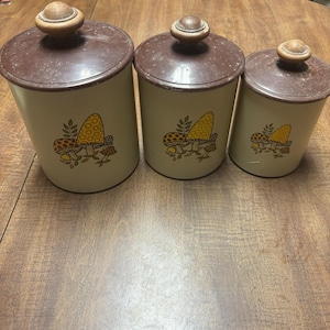 May include: A set of three vintage kitchen canisters. The canisters are off-white with brown lids and wooden knobs. Each canister features a mushroom design in shades of yellow, orange, and brown. The canisters are arranged in a row on a wooden surface.