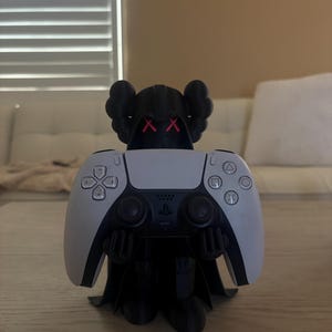 Op de afbeelding: Een zwart figuurtje met een capuchon, rode X-vormige ogen en uitgestrekte armen die een wit en zwart videogame-controller vasthoudt. De controller is voorzien van een richtingspad, twee analoge sticks en vier actieknoppen met geometrische symbolen.