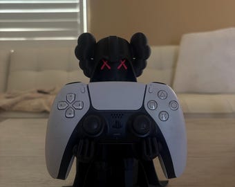 Könnte beinhalten: Eine schwarze Figur mit Kapuze, roten X-förmigen Augen und ausgestreckten Armen, die einen weiß-schwarzen Videospiel-Controller hält. Der Controller verfügt über ein Steuerkreuz, zwei Analogsticks und vier Aktionstasten mit geometrischen Symbolen.