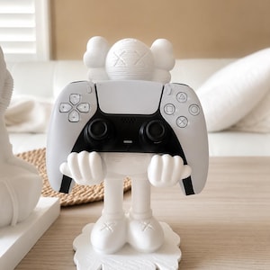 Op de afbeelding: Een wit mat beeldje met een cartoonachtig karakterontwerp met X-vormige ogen en gehandschoende handen die een zwart-witte videogamecontroller vasthouden. Het figuur staat op een ronde voet en ondersteunt de controller in een displaypositie.