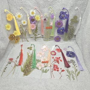 May include: Clear acrylic bookmarks adorned with dried flowers and colorful tassels. The bookmarks feature various floral arrangements, including daisies, purple flowers, and clovers. Some bookmarks have the word "sold" printed on them.