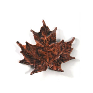 May include: A bronze-colored maple leaf decoration. The leaf has a textured surface and detailed veining, with a dark brown hue. The leaf is a decorative item, possibly for fall or autumn-themed decor.