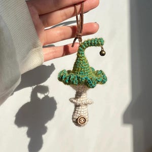 May include: A handmade crochet mushroom ornament with a green and blue cap, a beige stem, and a small wooden button. The ornament has a brown leather cord for hanging and a tiny bell attached to the cap.