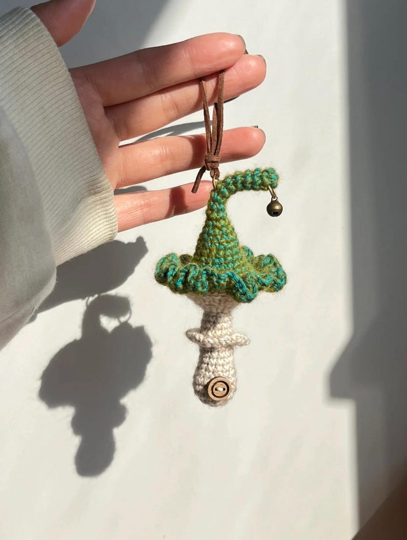 May include: A handmade crochet mushroom ornament with a green and blue cap, a beige stem, and a small wooden button. The ornament has a brown leather cord for hanging and a tiny bell attached to the cap.