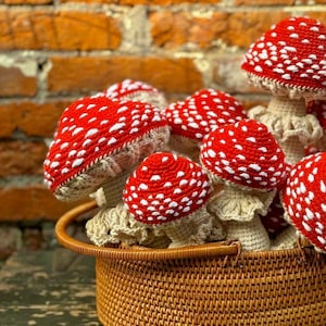 May include: A selection of handmade crochet mushrooms with red caps and white spots, presented in a woven basket. The mushrooms have beige stems, set against a brick wall background.