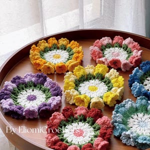 May include: A collection of handmade crochet flower coasters in various colors, including yellow, orange, pink, purple, red, and blue. Each coaster features a white center and a detailed petal design. The coasters are arranged on a wooden tray.