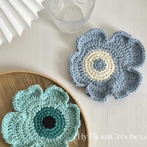 Puede incluir: Dos posavasos de flores de ganchillo hechos a mano. Uno es azul claro con un centro crema, el otro es verde azulado con un centro azul oscuro y negro. Los posavasos están sobre una bandeja de madera, con un vaso y un abanico de papel en el fondo.