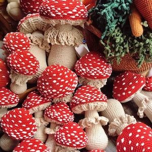 May include: A collection of handmade crochet mushrooms with red caps and white spots. The mushroom stems are beige. The image also shows a basket of crochet vegetables, including carrots and leafy greens. The mushrooms are arranged on a wooden surface.
