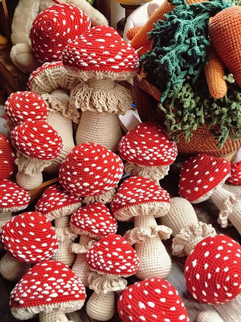 May include: A collection of handmade crochet mushrooms with red caps and white spots. The mushroom stems are beige. The image also shows a basket of crochet vegetables, including carrots and leafy greens. The mushrooms are arranged on a wooden surface.