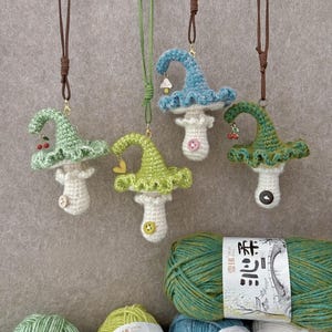 Può includere: Quattro ornamenti a forma di fungo all'uncinetto fatti a mano in tonalità di verde e blu, ognuno con un design unico e un piccolo ciondolo. I funghi sono sospesi da cordoncini marroni e verdi. Sotto sono visibili gomitoli di filato di vari colori.