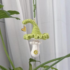 May include: A handmade crochet mushroom ornament with a green cap and white stem, adorned with a small yellow heart charm. The mushroom is suspended by a green cord, perfect for hanging. The ornament has a button detail on the stem.
