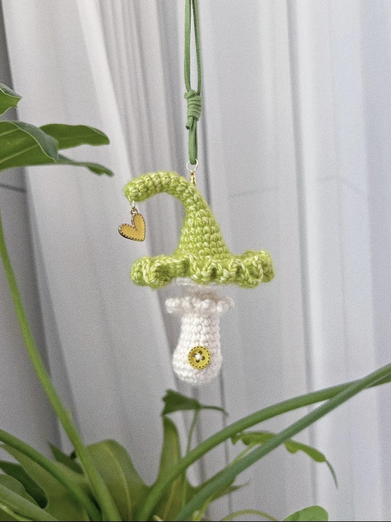May include: A handmade crochet mushroom ornament with a green cap and white stem, adorned with a small yellow heart charm. The mushroom is suspended by a green cord, perfect for hanging. The ornament has a button detail on the stem.