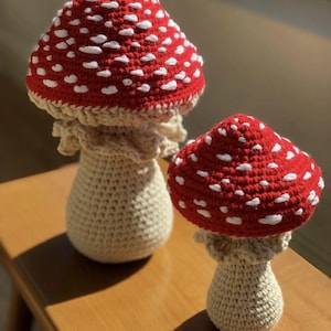 May include: Two crochet mushroom figurines. The mushroom caps are red with white spots, and the stems are beige. The mushrooms are sitting on a wooden surface. The larger mushroom is approximately 6 inches tall.