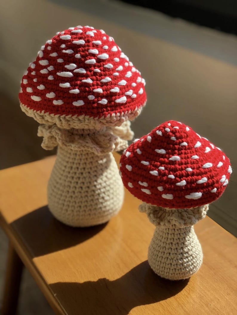 May include: Two crochet mushroom figurines. The mushroom caps are red with white spots, and the stems are beige. The mushrooms are sitting on a wooden surface. The larger mushroom is approximately 6 inches tall.